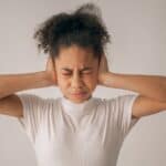 Despaired African American teenage girl with black hair and closed eyes covering ears while standing on white background in light studio