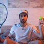 Sleepwalker with closed eyes holding tennis rocket and ball on bed