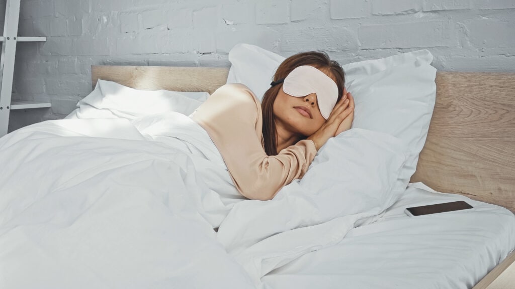 woman in eye mask sleeping near smartphone with blank screen